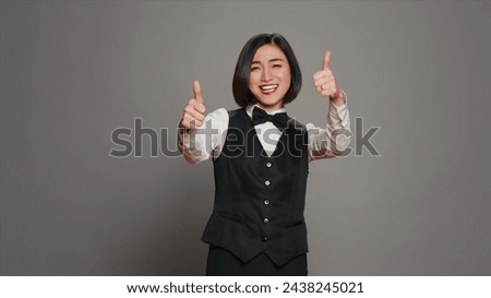 Asian reception staff doing thumbs up symbol on camera, showing approval and positive sign over grey background. Hotel receptionist giving like and okay gesture, showing agreement. Camera A.