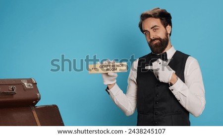 Elegant bellboy has conference room symbol in hand, showcasing wall pointer to lead hotel guests to all area on the premises. Experienced porter posing with an indicator wall sign.