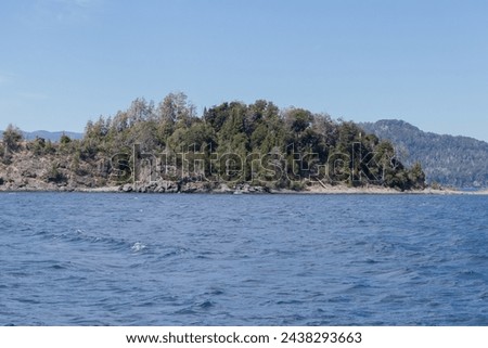 Horizontal photo of mountains full of vegetation and rocks, surrounded by tranquil waves of a lake's water, with a clear blue sky during a sunny day, in Lago Nahuel Huapi, Argentina
