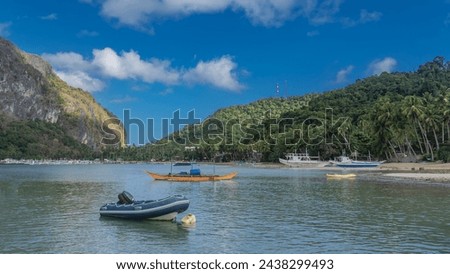 Inflatable, motorized, traditional Filipino bangka boats with double-outrigger dugout are anchored in the bay. Lush green vegetation, palm trees on the shore. A hill against a blue sky and clouds. Royalty-Free Stock Photo #2438299493