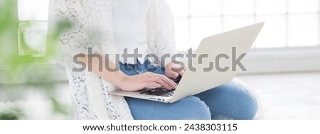 Faceless young woman in casual clothing sitting in living room operating laptop computer. There is a front blur of houseplants. Royalty-Free Stock Photo #2438303115
