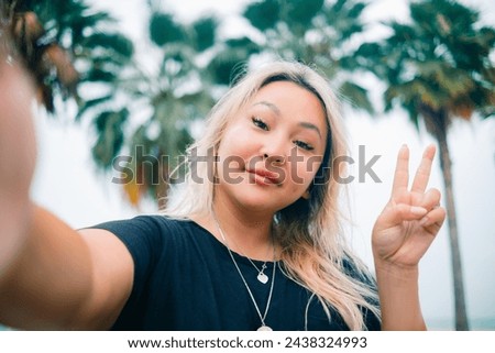 Young asian pretty woman shows V sign making selfie with happy smile with ocean beach palms on background. High quality photo