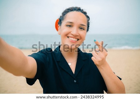 Young caucasian pretty woman with red dreadlocks shows heart sign making selfie with happy smile with ocean beach on background. High quality photo