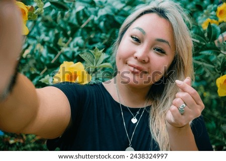 Young asian pretty woman shows heart sign making selfie with happy smile on background exotic plants. High quality photo