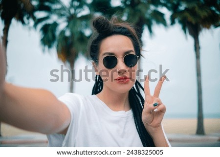 Young caucasian pretty woman shows V sign making selfie with happy smile with ocean beach palms on background. High quality photo