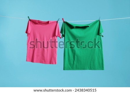 Two colorful t-shirts drying on washing line against light blue background Royalty-Free Stock Photo #2438340515