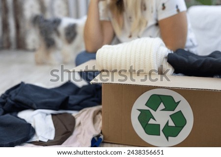 A box with a recycling sign with folded clothes stands on the floor. A woman sits in the background and goes through her wardrobe, sorting clothes. Selection of clothes for recycling.