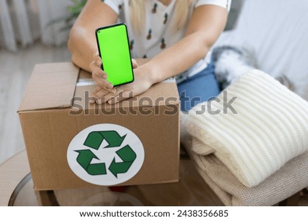 There is a box with a recycling sign on the table and stacked items next to it. A woman holds a phone with a green screen over a box. Advertising for recycling things.