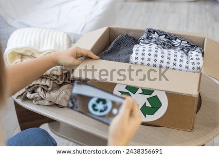 A recycling sign is on boxes of folded clothes that a woman is photographing on her phone. A woman wants to call the service and give her old clothes for recycling. Caring for nature.