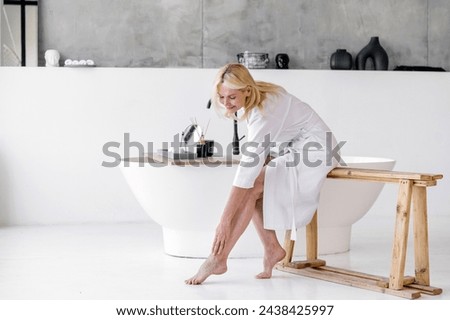 Mid age mature woman applying varicose prevention treatment cream massaging her barefoot legs sitting on bathtub wearing white bathrobe. Concept of health care and bodycare after menopause Royalty-Free Stock Photo #2438425997