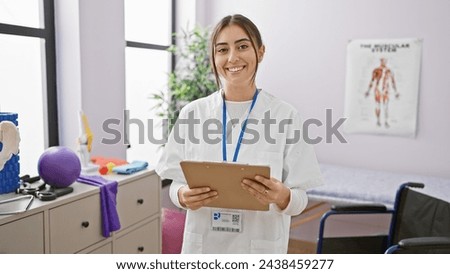 A smiling young hispanic woman in a white coat holding a clipboard, standing in a well-lit rehabilitation clinic room.