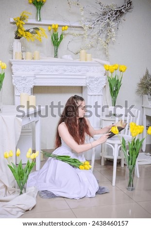 A beautiful young girl in a white dress and with long wavy hair reads a book against a background of yellow flowers.