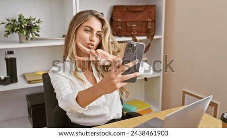 Bright young blonde woman worker, exuding confidence and positivity, enjoys successful business day. takes selfie picture with smartphone in elegant office interior, while working online on laptop.