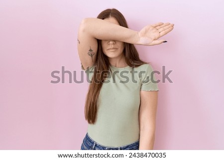 Beautiful brunette woman standing over pink background covering eyes with arm, looking serious and sad. sightless, hiding and rejection concept  Royalty-Free Stock Photo #2438470035