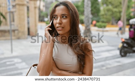 Stylish young hispanic woman engages in serious outdoor phone talk on sunlit city street Royalty-Free Stock Photo #2438470731