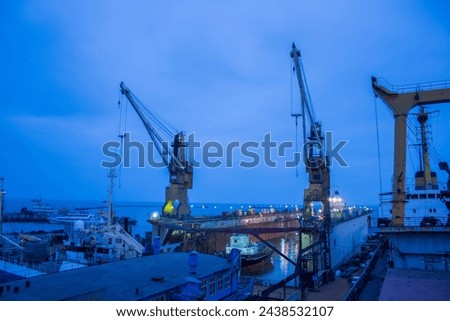 Twilight at shipyard with floating dock, vessels undergoing repair amid towering cranes. Maritime industry, heavy machinery at work, shipping business infrastructure, nautical vessel maintenance. Royalty-Free Stock Photo #2438532107