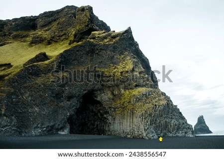 a man with a yellow jacket taking pictures of a random pattern of basalt rock formation in a mountain next to  a water body in southern region of Iceland near Vik