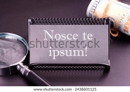 Latin proverb NOSCE TE IPSUM (know yourself) on the business card next to a roll of money and a magnifying glass on a black background