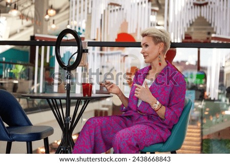 Beautiful mature blonde woman with a short haircut, an influencer, records a video blog while sitting at a table in a restaurant. Middle aged female blogger.