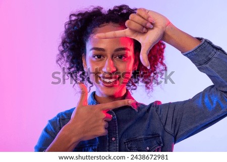 Cheerful black curly-haired woman creating frame with her fingers, playful and creative concept with gradient pink and blue neon background