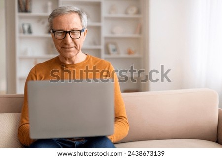 Seniors and digital world, modern technologies. Grandfather wearing eyeglasses sitting on sofa, using laptop computer at home, websurfing, reading news online, copy space Royalty-Free Stock Photo #2438673139