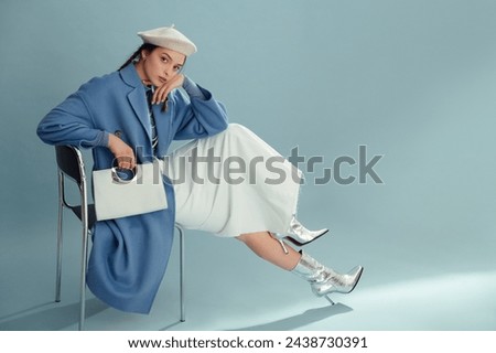 Fashionable confident woman wearing beret, elegant blue coat, white denim maxi skirt, silver ankle boots, holding stylish faux leather purse, posing on blue background. Full-length studio portrait Royalty-Free Stock Photo #2438730391