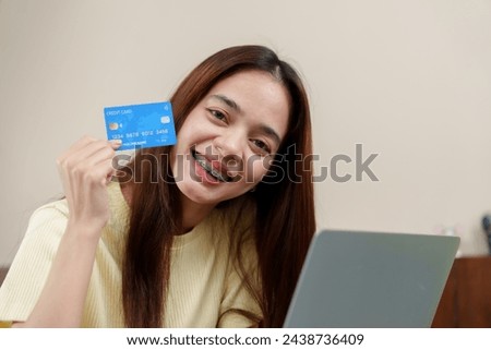 Content shopper orthodontic braces using credit card for an online purchase, seated with laptop. Young Asian woman beams, payment card in focus, partaking in the ease of online transactions at home.