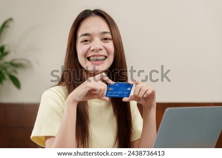 Ecstatic Asian young woman shopper with dental braces looks at laptop, holding credit card for an online purchase. Exuberant individual with a blue credit card smiles, anticipating an online buy
