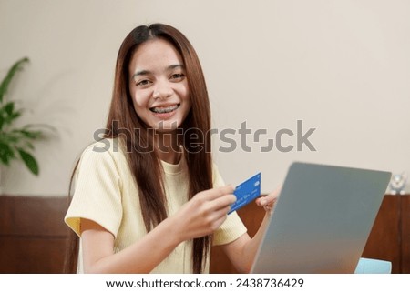 Jubilant Asian young woman with braces holds credit card, ready for online shopping spree, with laptop in front. Cheerful individual displays a bank card, preparing for e-commerce transaction,