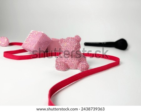 A sparkling pink teddy bear with shiny rhinestones, rest on a white background. A red strip intertwines around these playful objects, creating an aesthetic appeal.