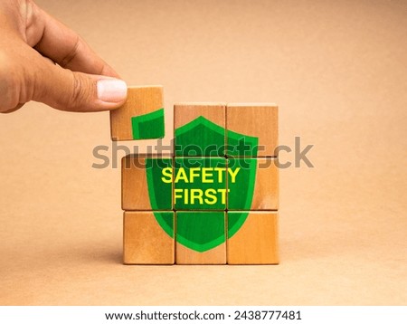 Safety first concept. Hand holding a last piece, put on the wooden cube block puzzle stack with text "SAFETY FIRST" on green shield icon isolated on brown background. Secure rules symbol at workplace.