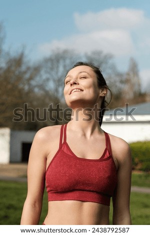 Smiling fitness female model posing relaxed in a park with eyes closed. She is wearing a red sport bra.