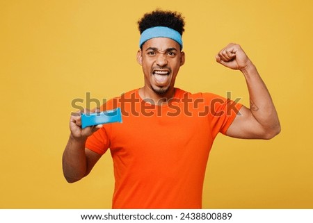 Young fitness trainer sporty man sportsman wear orange t-shirt hold energy bar protein snack show muscles scream training in home gym isolated on plain yellow background. Workout sport fit abs concept Royalty-Free Stock Photo #2438800889