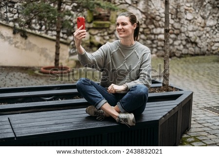 Happy 30s Women taking selfie and sitting on the bench outdoors. Young beautiful girl say Hi. Urban lifestyle concept, full body. High quality photo