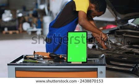 Mechanic in car service uses wrench to replace parts inside vehicle with green screen tablet in front. Expert utilizes professional tools to adjust malfunctioning automobile next to mockup device