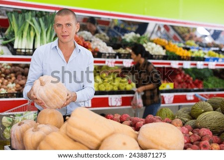Interested male shopper choosing ripe organic pumpkin while shopping in fruit and vegetable section of supermarket