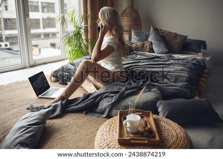 Blonde woman speaking phone working, making video call, watching movie on a laptop in cozy home bedroom modern interior. Home quarantine Covid-19 pandemic Corona virus. Remote work from home concept. Royalty-Free Stock Photo #2438874219