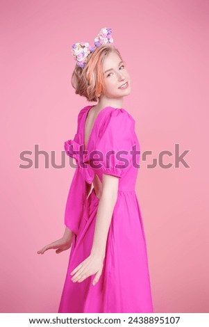 Kids and teenage fashion. A cute blonde teenage girl with a short haircut poses in a pink dress and a lovely headband with floral kitty ears. Pink background. Spring-summer look.