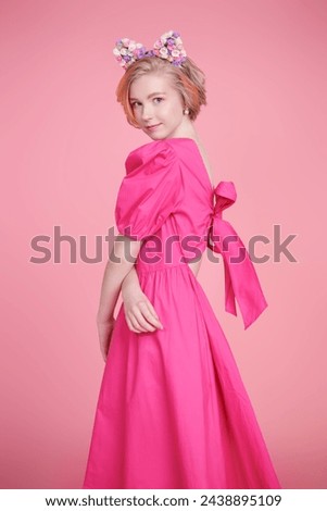 Spring-summer look. Kids and teenage fashion. A cute blonde teenage girl with a short haircut poses in a pink dress and a lovely headband with floral kitty ears. Pink background.
