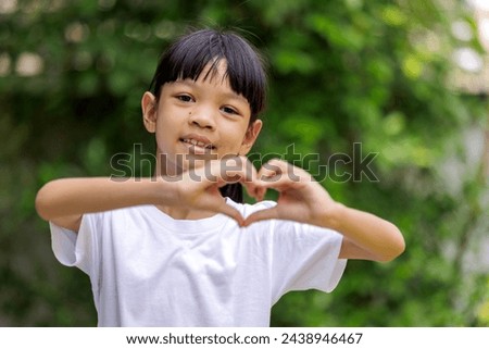 Portrait of a cute Asian girl. Wear a white t-shirt and standing with their hands together in the shape of a heart in different poses. She smiles brightly and naturally. Shady green garden background