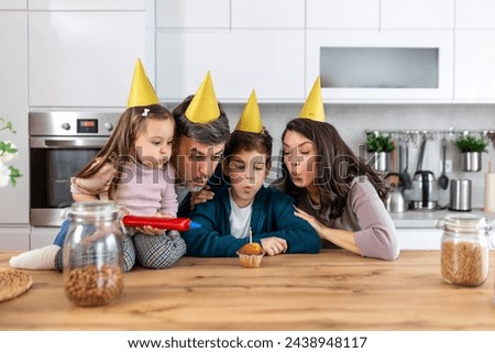 In the kitchen, a father blows out birthday candles on a cake, surrounded by his happy family. Laughter fills the air as they celebrate together, cherishing the moment of joy and togetherness.