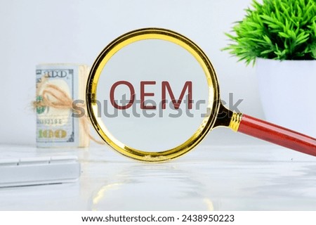 OEM original equipment manufacturer concept. Text through a magnifying glass on a gray background, a roll of banknotes and a green plant in the background