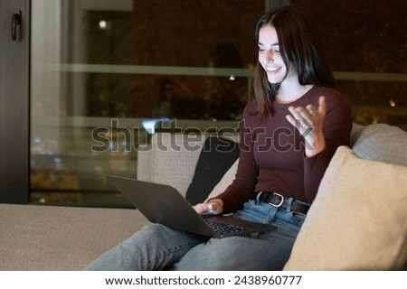 One cheerful woman having video call at home with friends sitting on sofa smiling at the laptop. Evening night time in window background. City life people modern lifestyle using online technology Royalty-Free Stock Photo #2438960777
