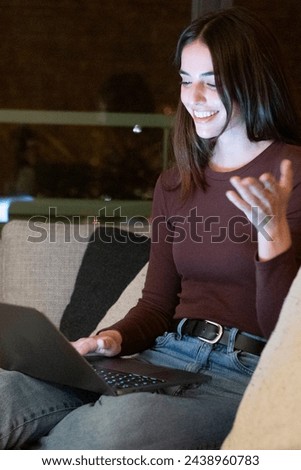 One cheerful woman having video call at home with friends sitting on sofa smiling at the laptop. Evening night time in window background. City life people modern lifestyle using online technology Royalty-Free Stock Photo #2438960783