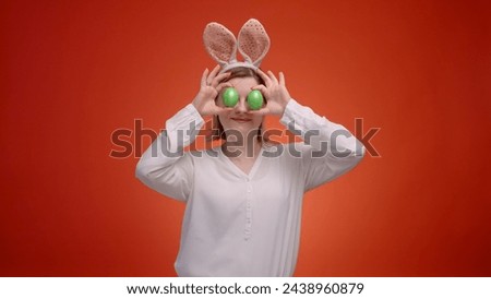 Woman in Easter bunny ears with colored eggs dancing on a red background and putting eggs on eyes, the concept of coming Easter and preparing for celebration