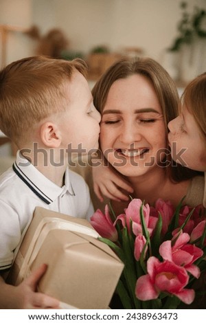 Daughter and little son kissing and hugging mom and make surprise for Mothers Day. Children give bouquet of tulip flowers. Childs gives present box congratulates surprised mom. Happy family closeup