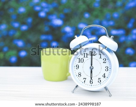 Front view of white vintage alarm clock and green cup of coffee on the table in the garden with purple flowers background.