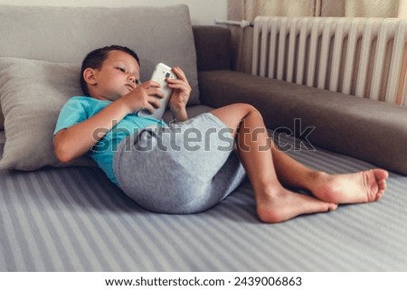 Portrait of a cute little child looking at modern mobile while lying on cozy bed in room. Wistful boy using gadget concept.