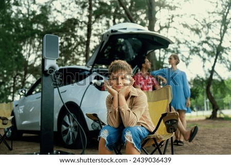 Little boy portrait sitting on camping chair with his family in background. Road trip travel with alternative energy charging station for eco-friendly car concept. Perpetual