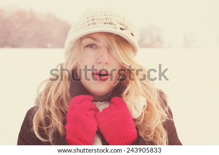 winter woman in snow photo, looking and blows breath at camera outside on cold winter vintage day. Portrait Caucasian female model with pink gloves and white hat with scarf in first snow
